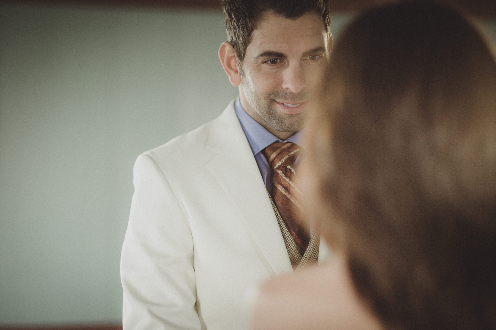 A man in white suit and tie looking at his reflection.